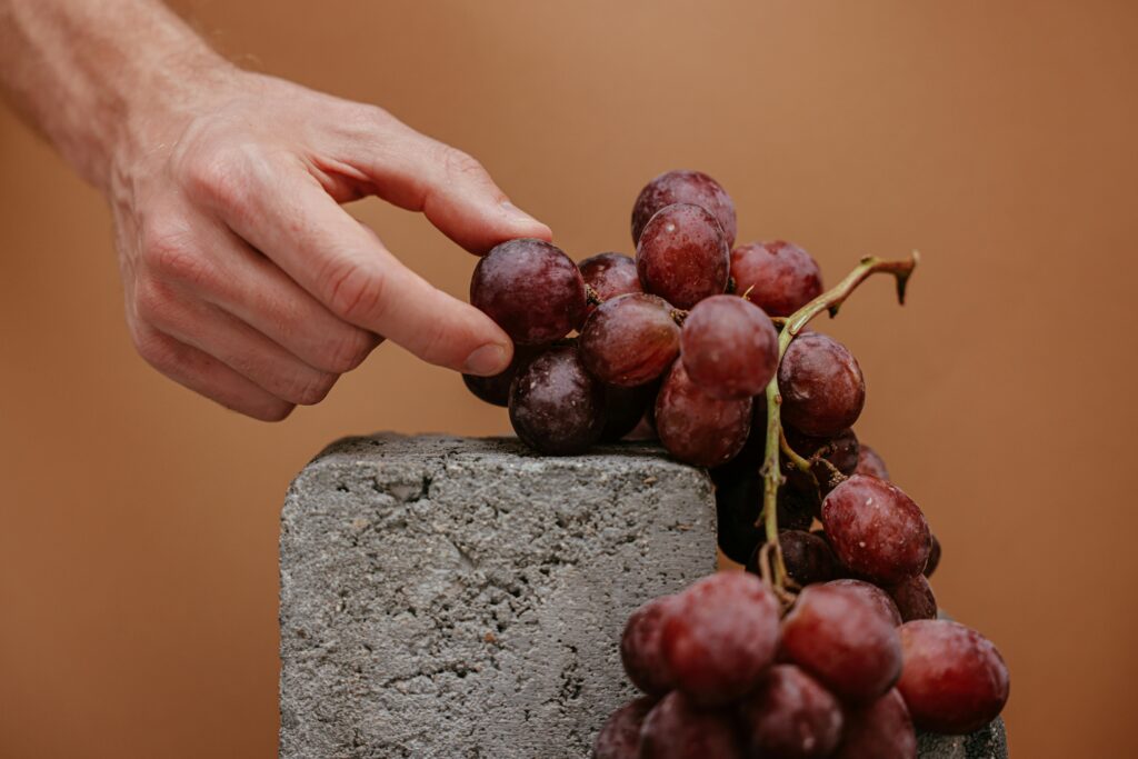 A close-up shot of red grapes and a hand on a stone block against a brown background.