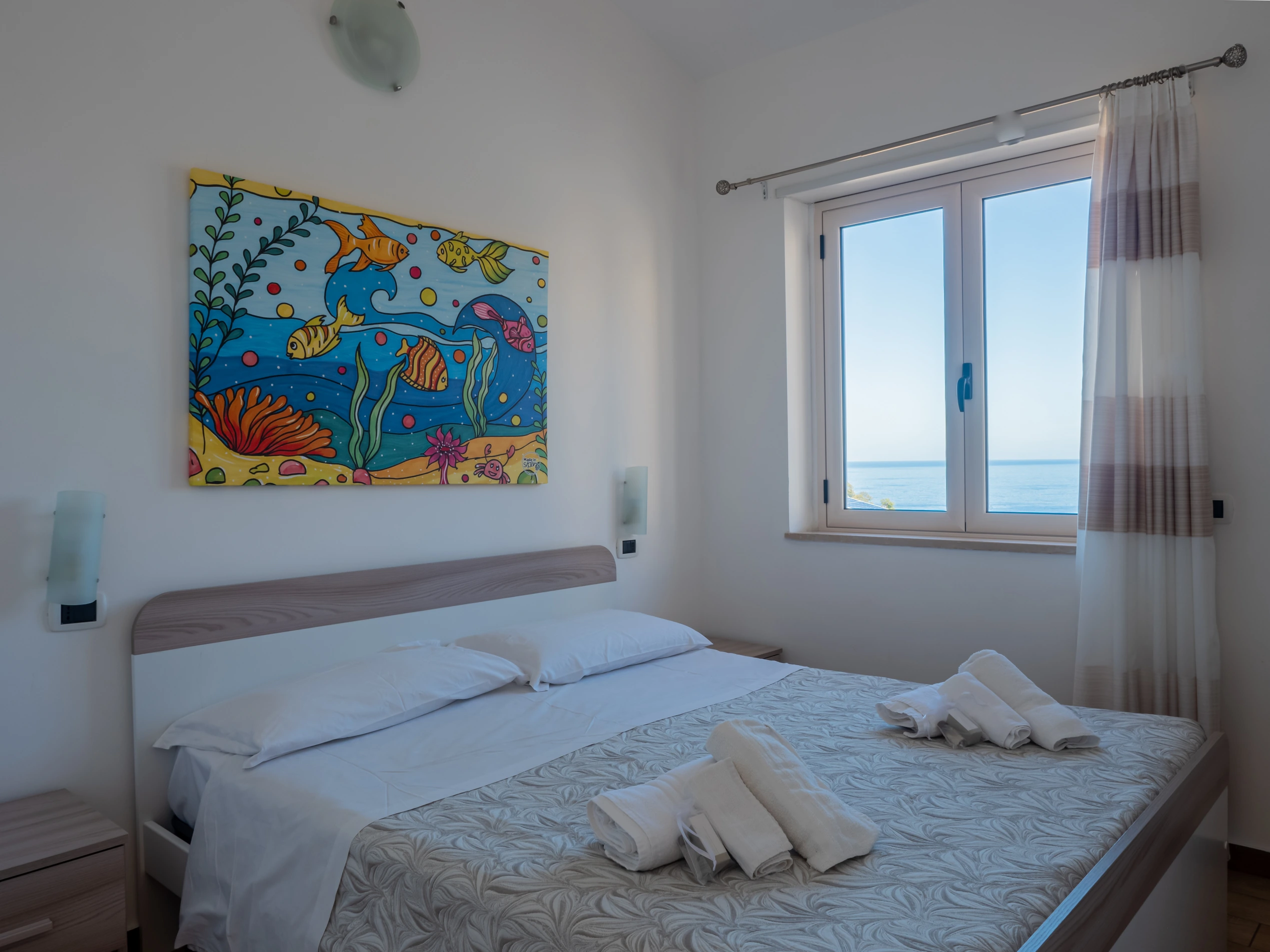 Camera da letto matrimoniale del bilocale all'Airone Residence Zambrone con letto pronto, asciugamani bianchi, quadro marino colorato e finestra con vista mare.