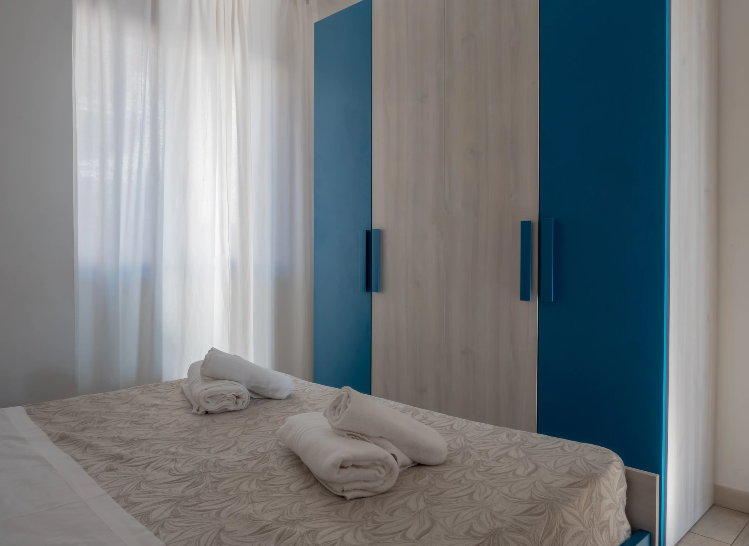 Camera matrimoniale con asciugamani puliti sul letto e grande armadio moderno color legno e blu mare all'Airone Residence Zambrone.