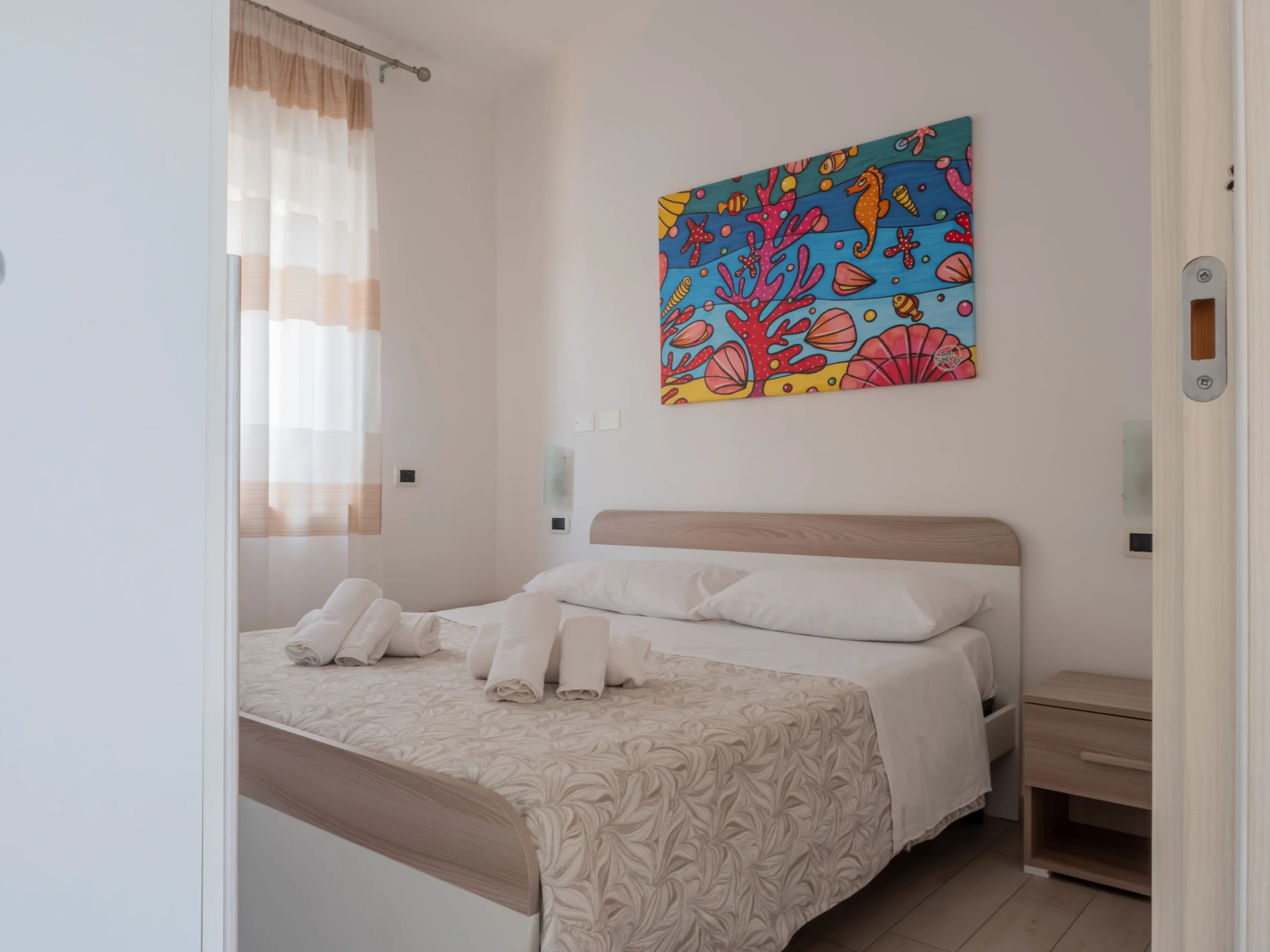 Camera da letto matrimoniale del bilocale all'Airone Residence Zambrone con asciugamani bianchi sul letto, comodino e quadro colorato con coralli.