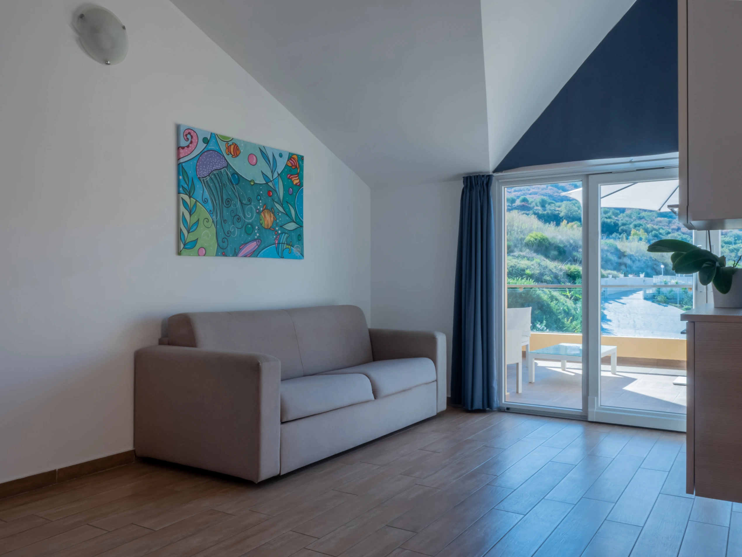 Moderno divano in tessuto beige nel soggiorno di un appartamento all'Airone Residence Zambrone, con quadro marino e porta finestra aperta sul terrazzo.