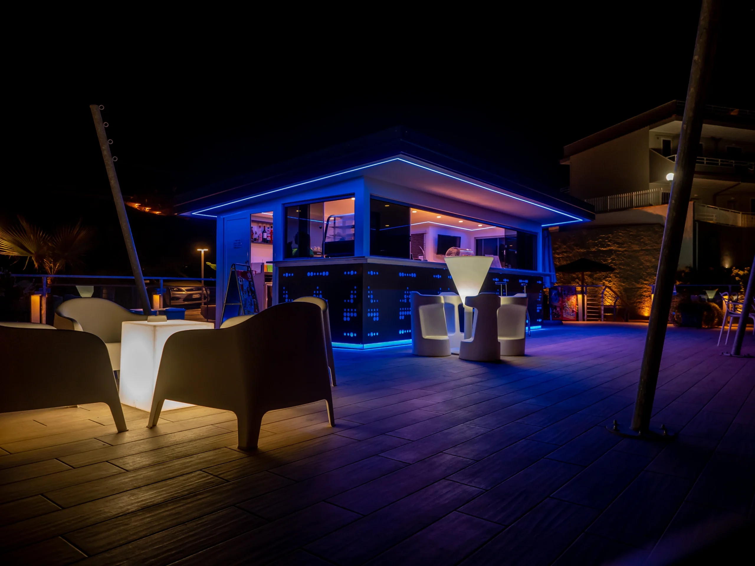 Il cocktail lounge bar all'aperto dell'Airone Residence Zambrone di notte con illuminazione a LED blu e tavoli luminosi.