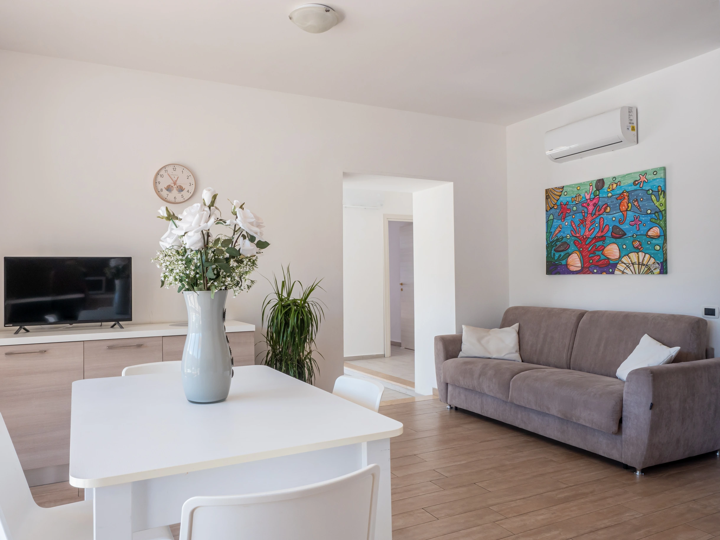 Soggiorno climatizzato dell'Airone Residence Zambrone con tavolo da pranzo bianco, divano, quadro moderno e mobile TV.