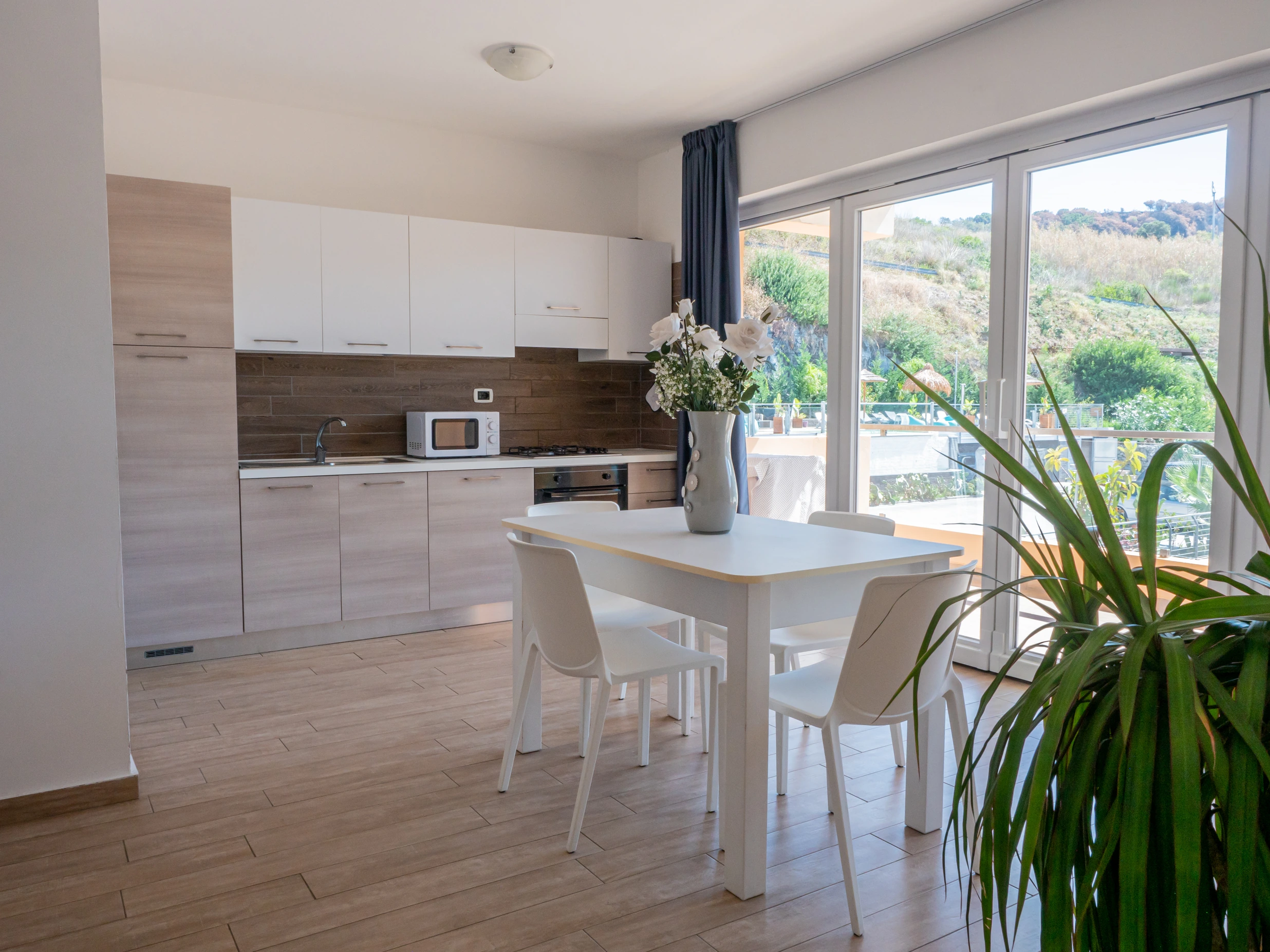 Zona giorno open space molto luminosa con cucina lineare moderna, tavolo da pranzo bianco e grande vetrata scorrevole all'Airone Residence Zambrone.