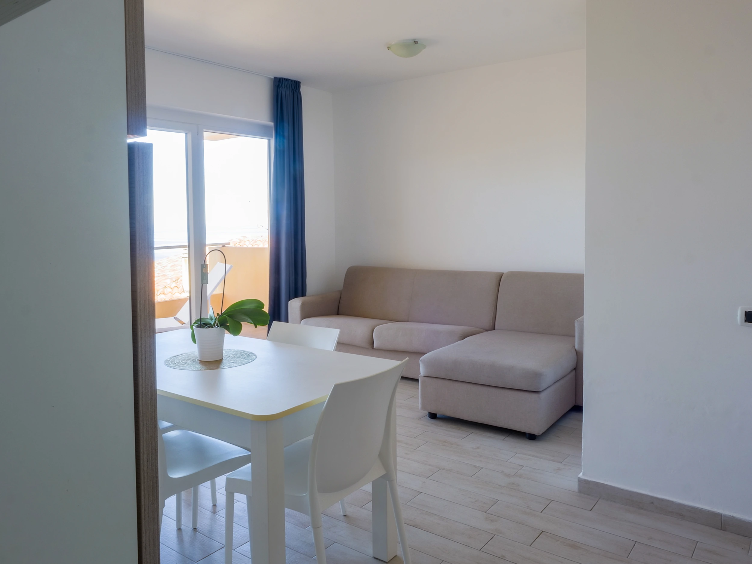 Soggiorno luminoso di un appartamento all'Airone Residence Zambrone con divano ad angolo beige, tavolo da pranzo bianco e porta finestra sul terrazzo.