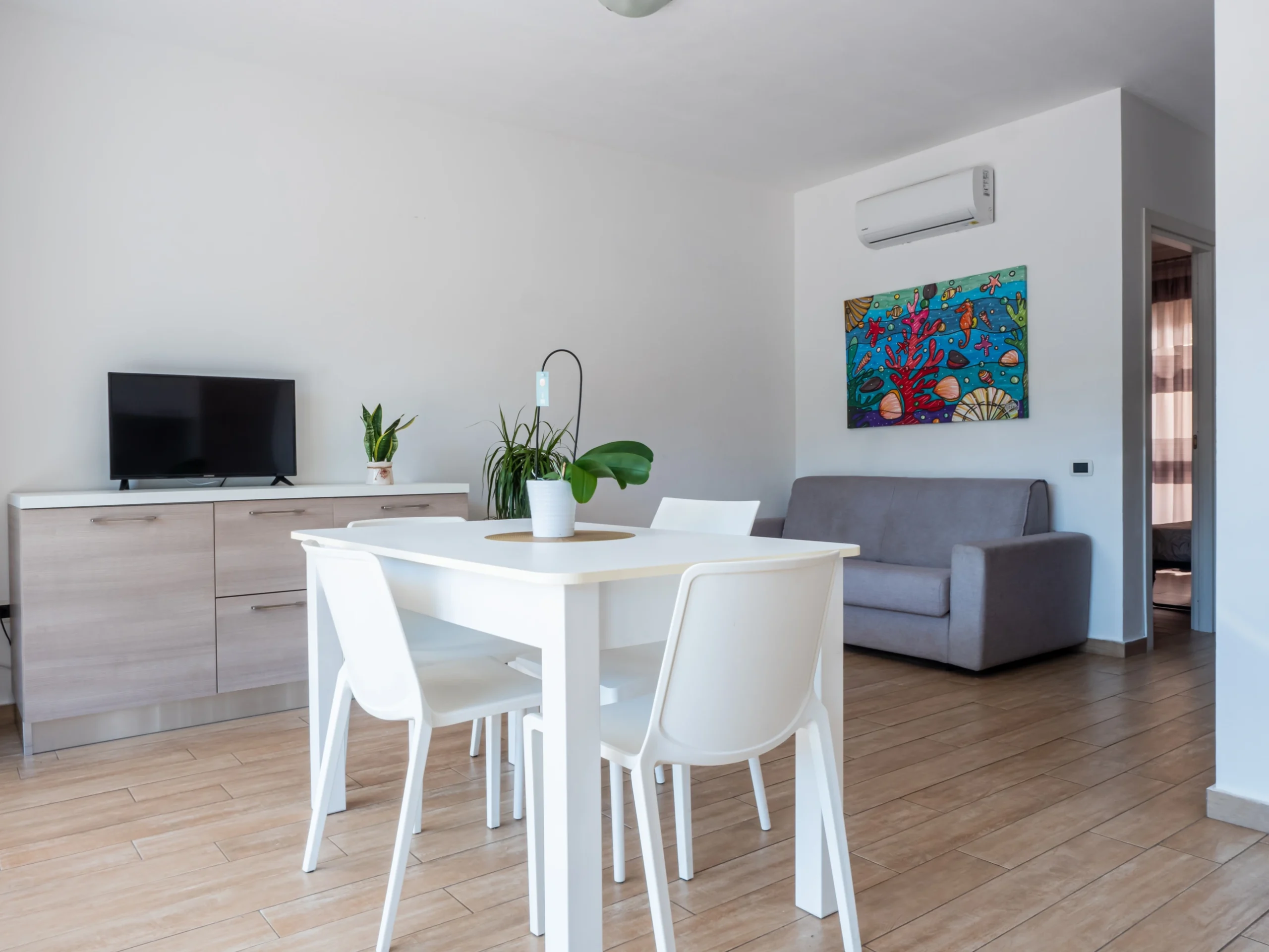 Tavolo da pranzo bianco con sedie, mobile TV con schermo piatto e divano beige sotto un quadro marino colorato nel soggiorno dell'Airone Residence Zambrone.