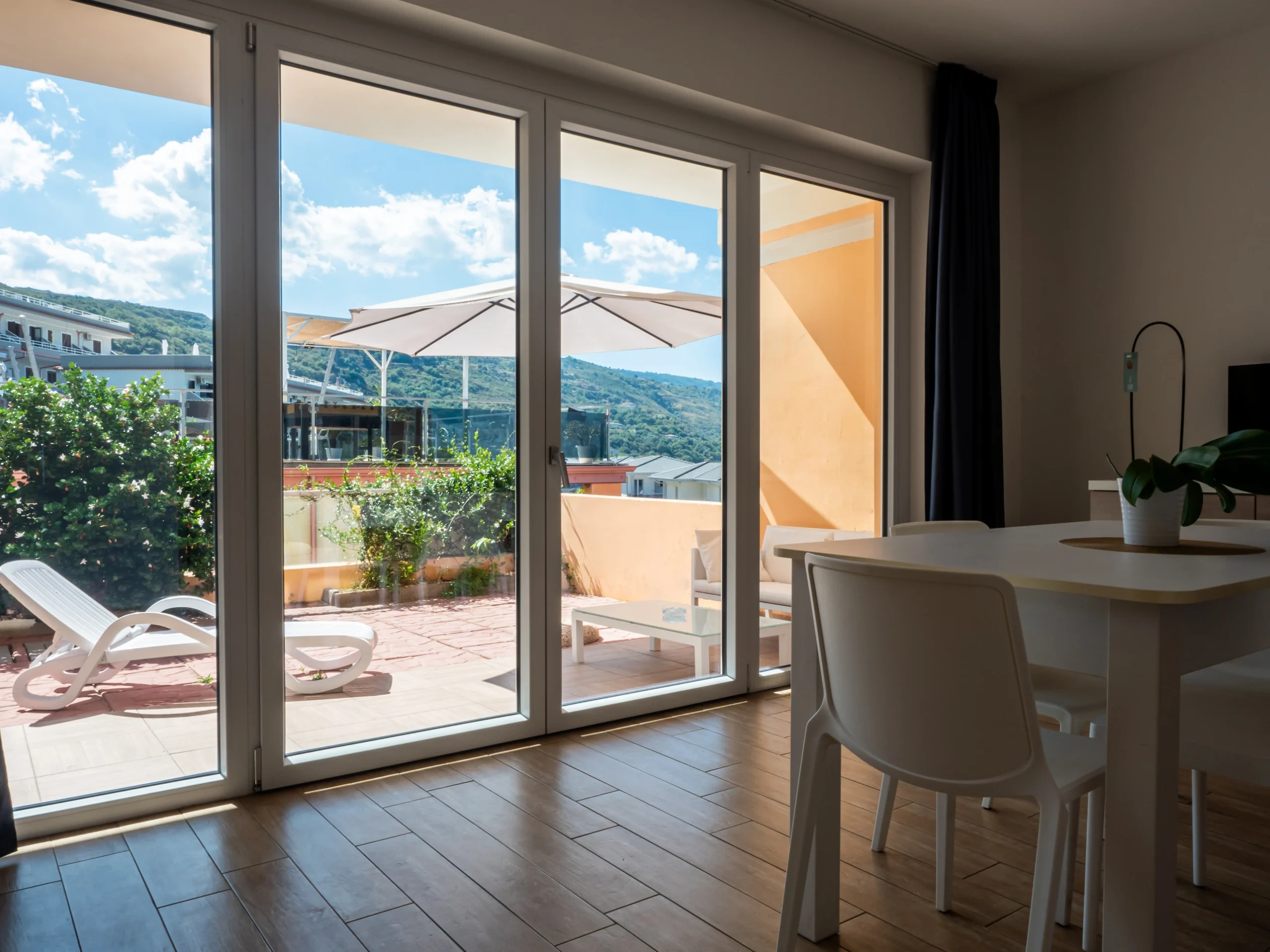 Interno del soggiorno con tavolo da pranzo bianco e ampia vetrata aperta sul terrazzo solarium attrezzato con lettino e ombrellone all'Airone Residence Zambrone.