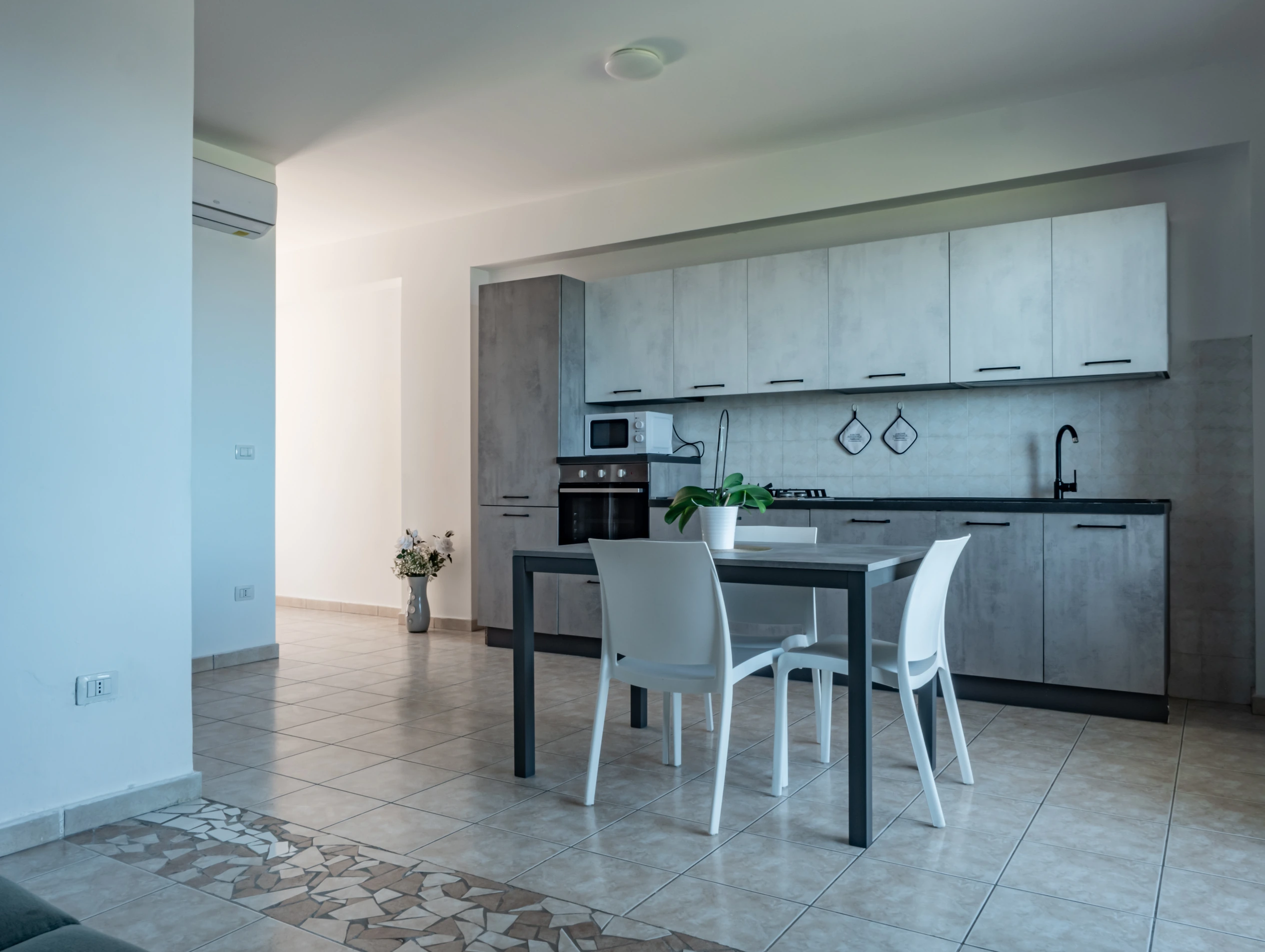 Ampio soggiorno open space del bilocale standard dell'Airone Residence Zambrone con cucina moderna effetto cemento grigio, tavolo da pranzo e climatizzatore.