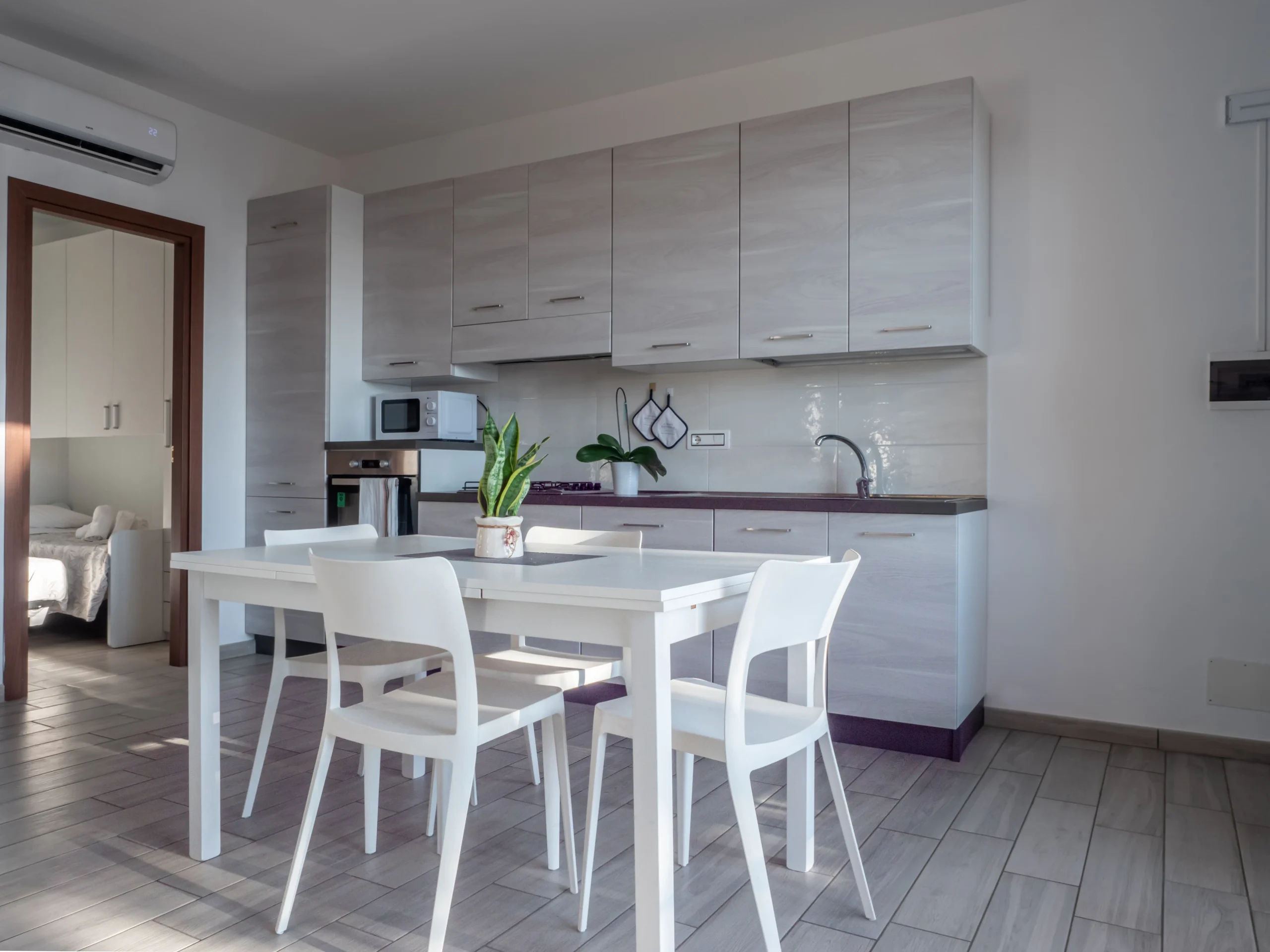 Tavolo da pranzo bianco con sedie, cucina lineare attrezzata in legno chiaro e porta aperta sulla camera da letto all'Airone Residence Zambrone.