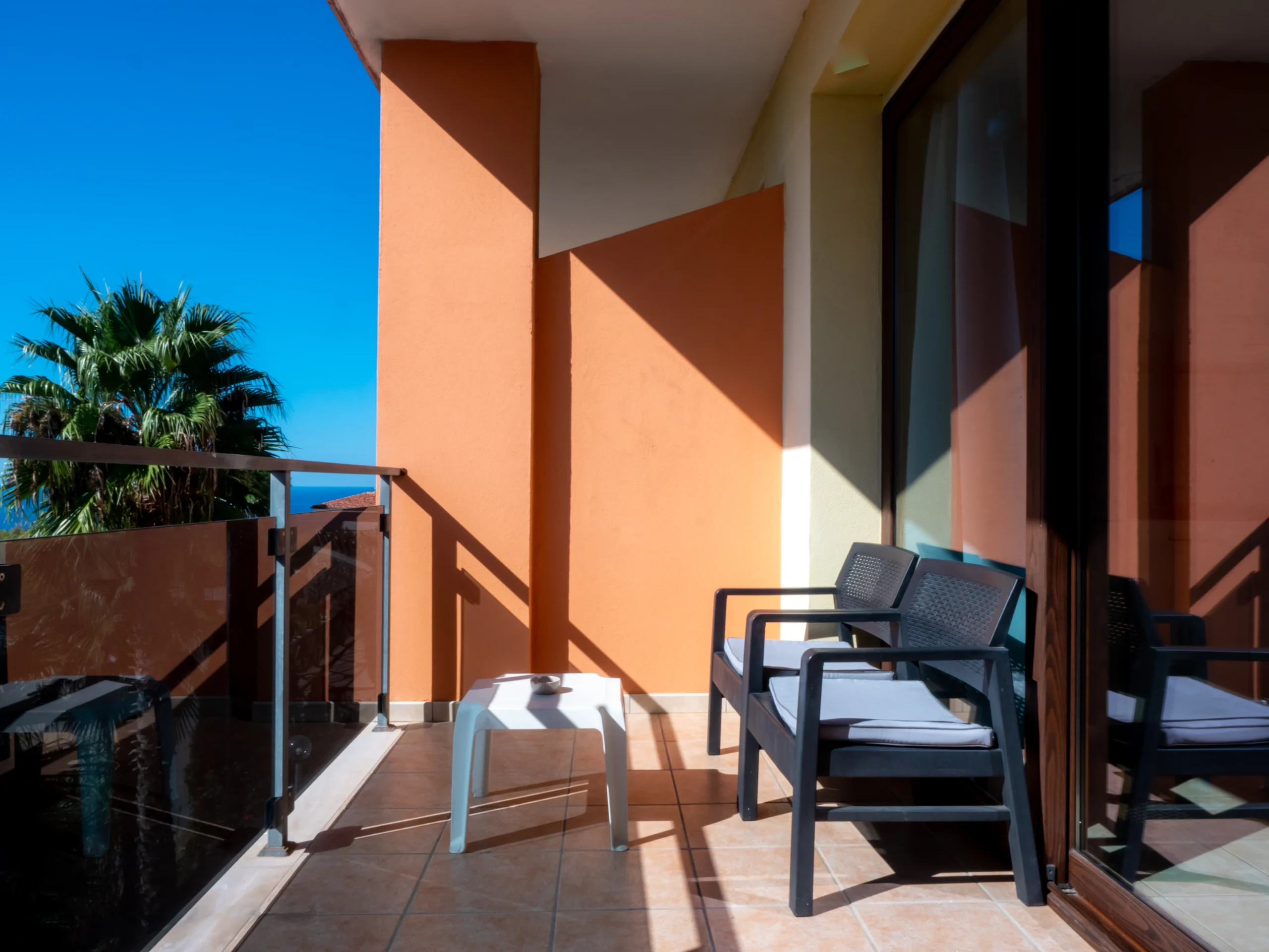 Terrazzo coperto privato all'Airone Residence Zambrone argettao con poltrone da esterno scure, cuscini grigi, tavolino bianco e vista su una palma.