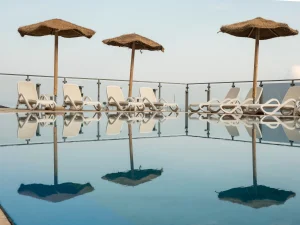 Piscina a sfioro all'Airone Residence Zambrone con lettini bianchi e ombrelloni in paglia riflessi sull'acqua, con vista mare sullo sfondo.